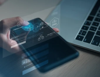 Eine Hand hält ein Smartphone, auf dem holografische Cybersecurity-Icons erscheinen; rechts Laptop auf Holztisch.