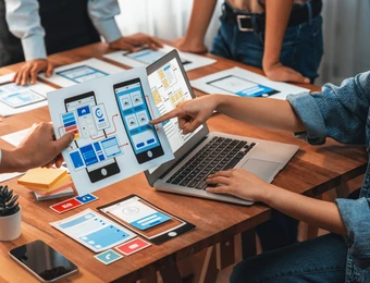 Mehrere Personen um einen Tisch; Laptop geöffnet, Tablet mit App-Layout, Smartphone-Mockups und Karten auf dem Tisch.