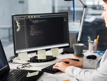 Person sitzt an Schreibtisch mit Monitoren; zentraler Monitor zeigt Quellcode, Laptop daneben; Tasse und Stifte.