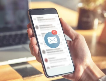 Eine Hand hält ein Smartphone mit einer E-Mail-Benachrichtigung (12); im Hintergrund ein Laptop.
