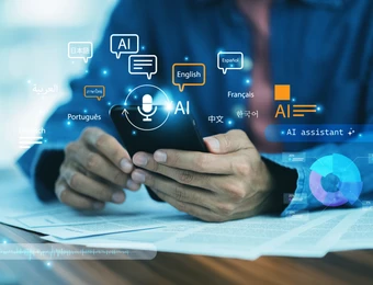 Person hält Smartphone; holografische AI-Icons, Sprachen-Symbole, Diagramme über einem Tisch mit Papieren.
