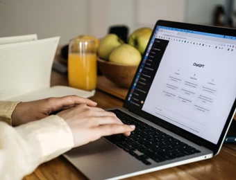 Person tippt auf einem silbernen Laptop an einem Holztisch; im Hintergrund Obstschale und Glas Orangensaft.