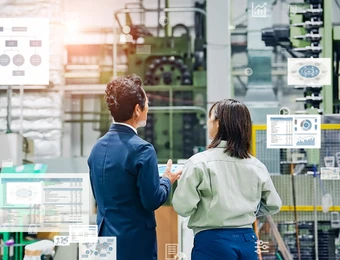 Zwei Personen stehen in einer Industrieanlage vor Maschinen, umgeben von Monitoren und Diagrammen.