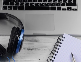 Top-Down: Laptop, Kopfhörer mit blauen Akzenten, Kabel, Spiralnotizbuch mit Stift, Brille, Pflanze im Topf.