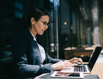 Eine Frau mit Brille arbeitet an einem Laptop an einem Holztisch draußen, Smartphone daneben.