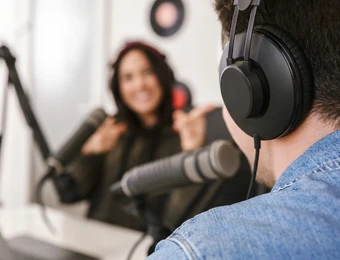 Zwei Personen in einem Podcast-Studio; Vordergrund: Person mit Kopfhörern, Rücken sichtbar; Hintergrund: Frau am Mikrofon.