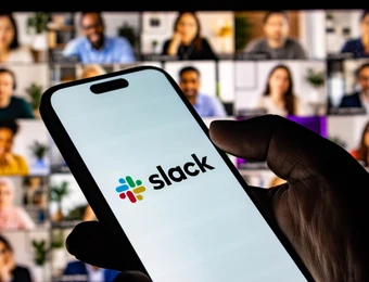 Nahaufnahme: Hand hält Smartphone mit Slack-Logo, Hintergrund aus vielen Video-Konferenzfenstern.