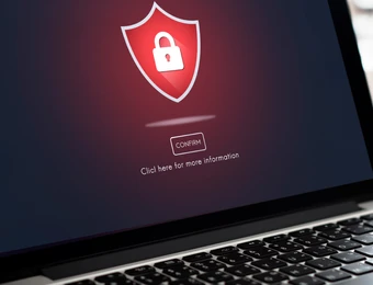 Laptop auf Holztisch, Bildschirm zeigt rotes Schutzschild mit Schloss; Glas im Hintergrund.