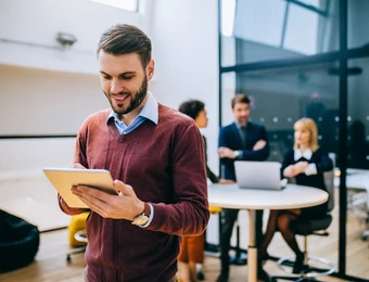 Mann mit Bart im burgunderroten Pullover hält Tablet; im Hintergrund arbeiten Personen an einem runden Tisch mit Laptop.