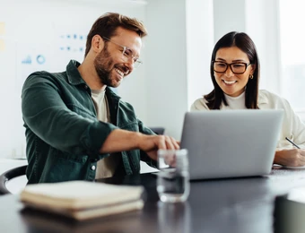 Zwei Personen sitzen an einem Tisch mit Laptop; Mann links lächelt, Frau rechts schreibt; Glas Wasser und Notizbuch sichtbar.