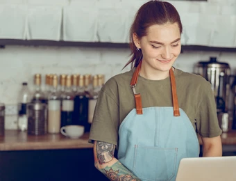 Person mit hellblauer Schürze hinter einer Café-Theke, lächelt.