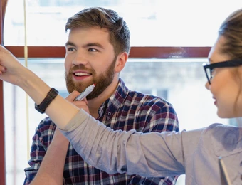 Zwei Personen in einem Büro schauen auf eine hängende Reihe bunter Notizen; Mann lächelt, Frau zeigt darauf.