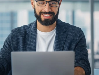 Ein Mann mit Brille und Bart sitzt mit offenem Laptop in einem Büro; im Hintergrund Glasfenster und bunte Klebezettel.