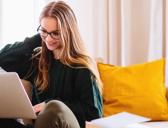 Eine Frau sitzt auf einer hellen Couch und schaut auf einen Laptop; gelbes Kissen, Kamera rechts.