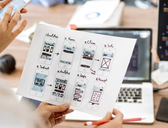 Person hält UI-Entwürfe auf Papier; Laptop mit Code im Hintergrund; Smartphone und Notizbuch auf dem Tisch.