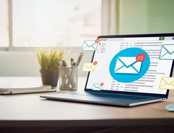 Laptop auf Holztisch; Bildschirm zeigt E-Mail-Symbol mit Benachrichtigungen, daneben Notizbuch, Pflanze und Stifte; Fenster im Hintergrund.