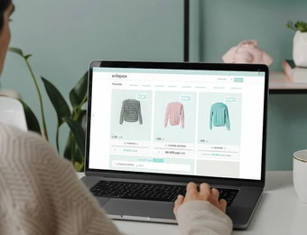 Person von hinten sitzt vor Laptop; Bildschirm zeigt drei Pullover-Modelle in Grau, Rosa und Mintgrün.