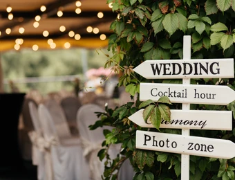 Weißes Holzschild mit Pfeilen (WEDDING, Cocktail hour, Photo zone) an einer Grünpflanze; im Hintergrund Tische und Lichter.