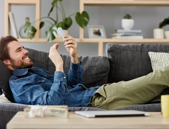 Mann liegt auf grauer Couch, schaut aufs Smartphone; Wohnzimmer mit Pflanzen, Regal und gelber Tasse.