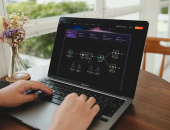 MacBook auf Holztisch, Hände tippen; Vase mit Blumen links, helles Fenster im Hintergrund; Bildschirm zeigt Diagramm.