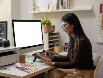 Frau sitzt am Schreibtisch mit Grafiktablett, Stift in der Hand; Monitor weiß, Farbmuster und Kamera auf dem Tisch.