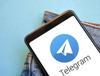 Smartphone mit Telegram-Logo (weißer Papierflieger auf blauem Kreis) auf blauem Hintergrund neben Jeansstoff.
