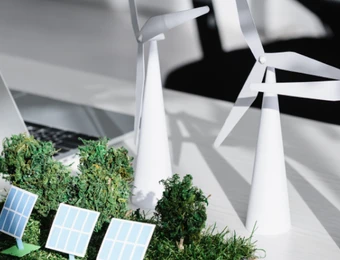 Laptop auf Tisch, weiße Windräder im Hintergrund, Solarzellen-Module und grüne Pflanzen im Vordergrund.