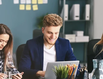 Vier Personen sitzen an einem Tisch mit Laptops; eine Pflanze in der Mitte, Hintergrund mit Haftnotizen.