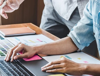 Zwei Personen arbeiten: Eine Person tippt auf Laptop, die andere zeigt mit Stift auf Farbpaletten und Notizblock.