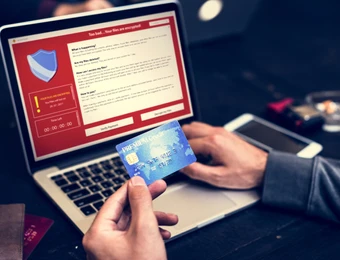 Person hält eine blaue Kreditkarte vor einem Laptop mit rotem Display und Schild-Symbol; Textfelder sichtbar.
