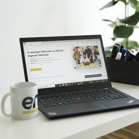 Offener Laptop auf weißem Tisch, Bildschirm zeigt Website; weiße Tasse links, Pflanze rechts im Hintergrund.
