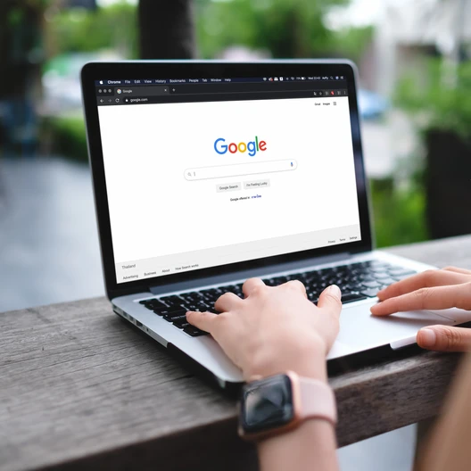 Person tippt auf einem Laptop auf einem Holztisch im Freien; Google-Startseite auf dem Bildschirm sichtbar.