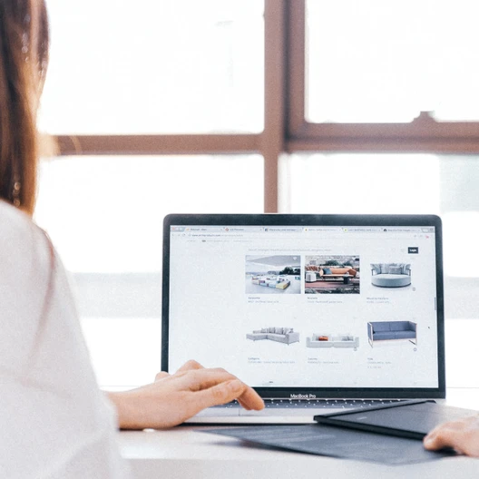 Zwei Personen sitzen an einem Tisch und arbeiten an einem Laptop; der Bildschirm zeigt eine Webseite, im Hintergrund Fenster.