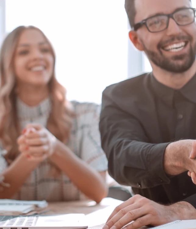 Gruppe von Büroangestellten; zwei schütteln sich die Hände, eine lächelt; Laptop, Pflanze und Fenster im Hintergrund.