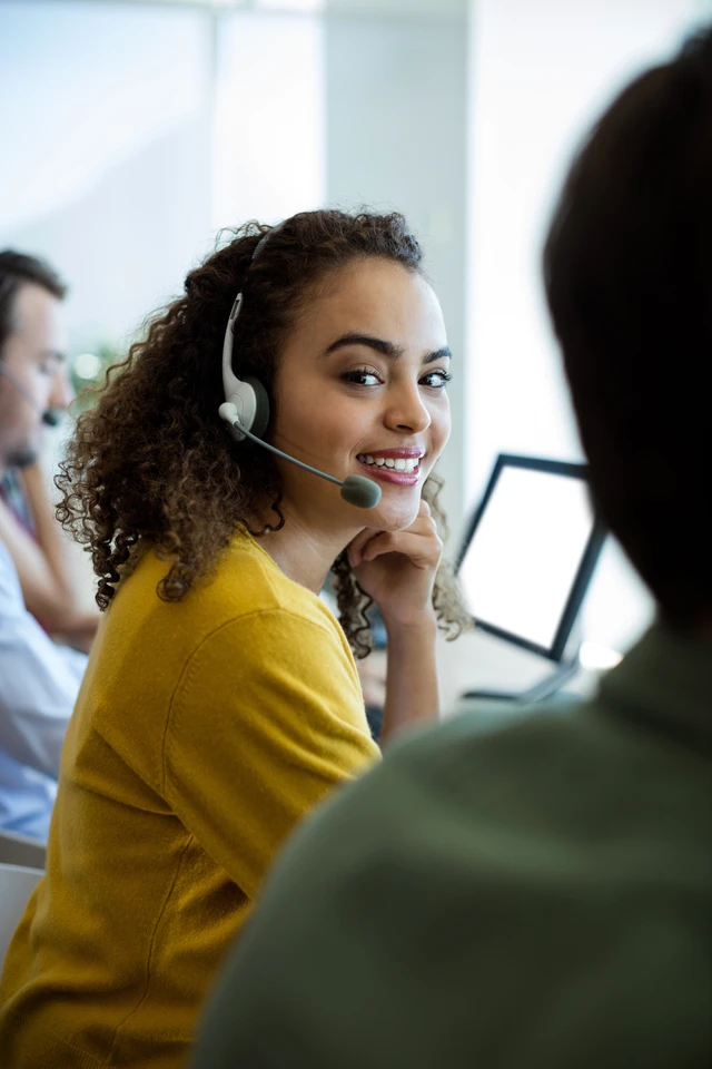 Frau mit lockigem Haar, gelbem Oberteil, Headset mit Mikrofon, lächelnd vor Monitoren in einem Büro.