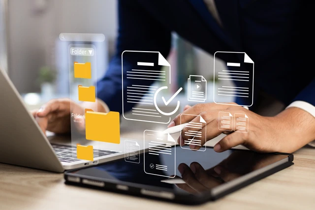 Person nutzt Laptop und Tablet; gelbe Ordner-Icons und schwebende Dokumente mit Häkchen erscheinen über dem Bildschirm.