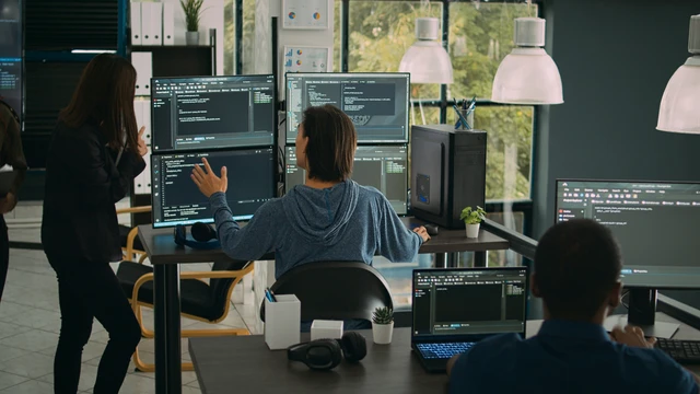 Drei Personen arbeiten in einem Büro; mehrere Monitore zeigen Code; zentrale Person sitzt und zeigt auf einen Bildschirm.