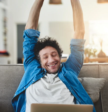 Ein Mann sitzt auf einer Couch, Arme über dem Kopf verschränkt, lächelt, vor Laptop.