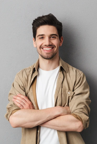Junger Mann mit beigem Hemd über weißem T‑Shirt, Arme verschränkt, lächelnd vor grauer Wand.
