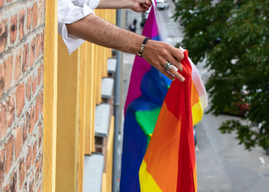 Arm einer Person mit Schmuck hält eine Regenbogenflagge neben gelb gestrichener Fensterlaibung an einer Backsteinwand.