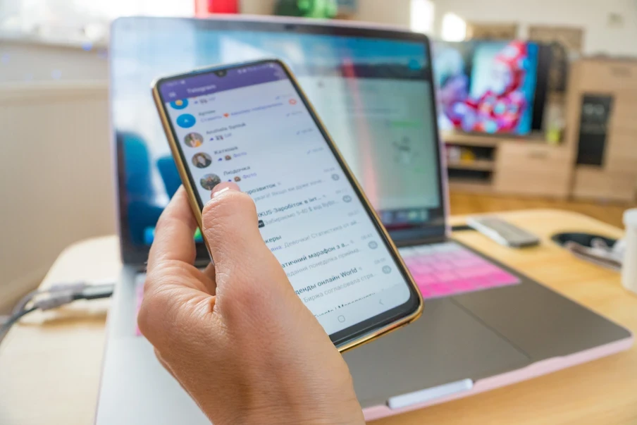 Eine Hand hält ein Smartphone mit Chat-App; im Hintergrund ein Laptop auf dem Schreibtisch.