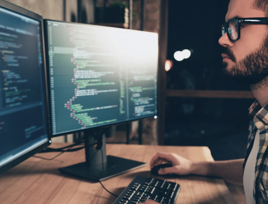 Mann mit Bart und Brille sitzt vor zwei Monitoren mit Code, Tastatur vor ihm, dunkler Hintergrund.