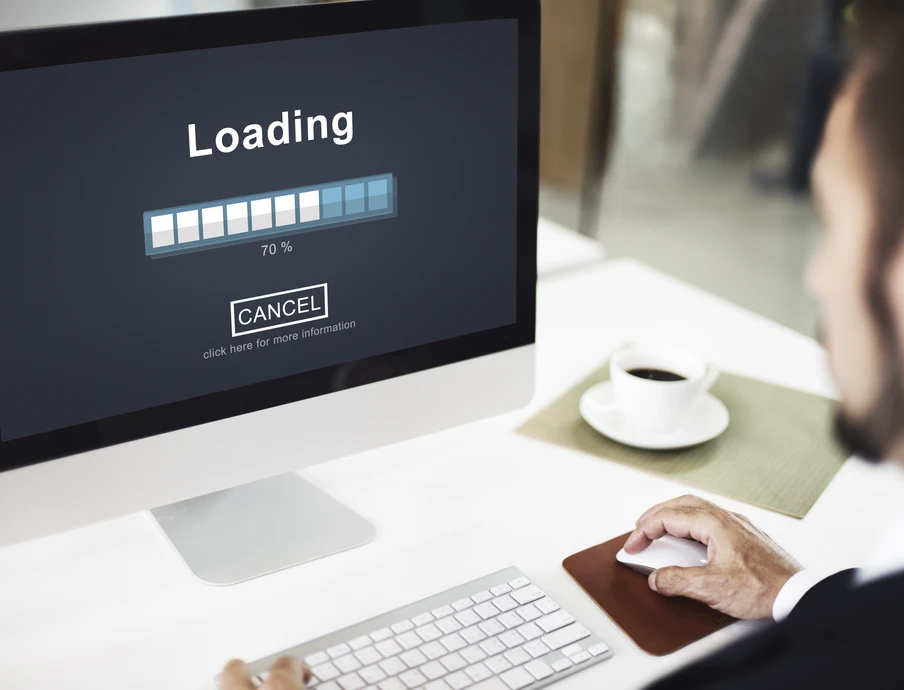 Monitor zeigt Ladebildschirm mit Fortschrittsbalken; ein Mann sitzt am Schreibtisch neben Tastatur und Kaffeetasse.