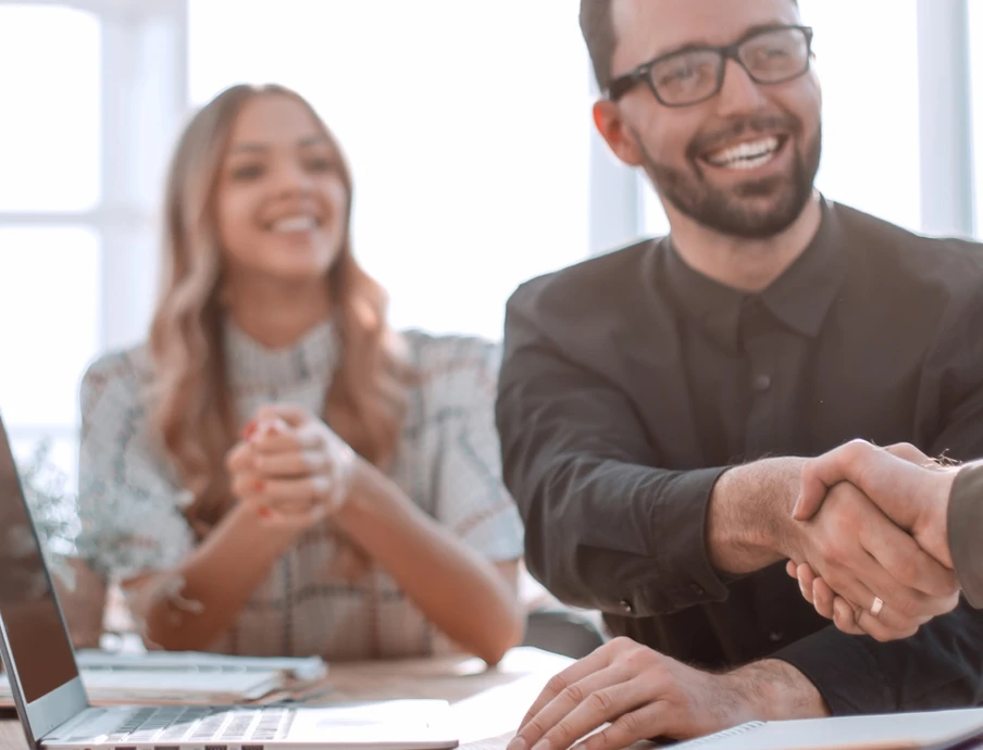 Gruppe von Büroangestellten; zwei schütteln sich die Hände, eine lächelt; Laptop, Pflanze und Fenster im Hintergrund.