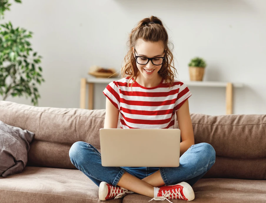 Frau sitzt auf dem Sofa, nutzt Laptop; rotes gestreiftes Oberteil, Jeans, Brille; Pflanze und Regal im Hintergrund.