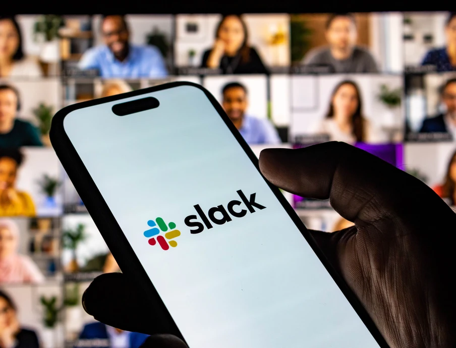 Nahaufnahme: Hand hält Smartphone mit Slack-Logo, Hintergrund aus vielen Video-Konferenzfenstern.