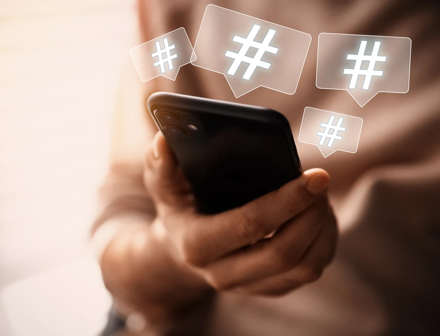 Person hält Smartphone, umgeben von schwebenden Hashtag-Symbolen in quadratischen Blasen.