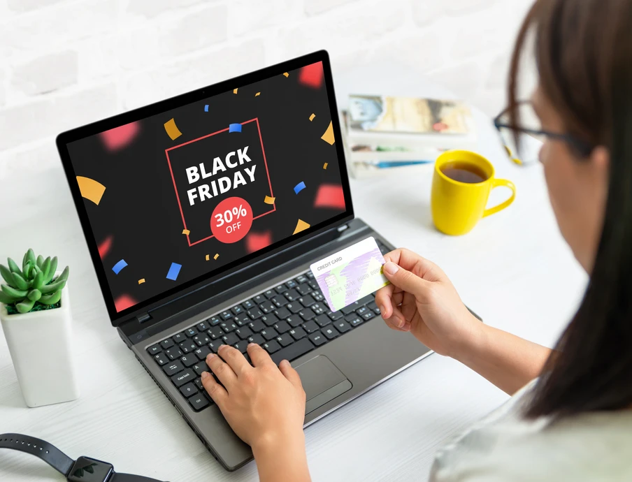 Person nutzt Laptop; Bildschirm zeigt Black Friday 30% Off. Hand hält Kreditkarte; gelbe Tasse, Pflanze auf weißem Tisch.