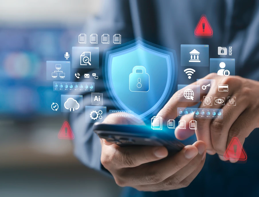 Eine Hand hält ein Smartphone; holografische Sicherheits-Icons und ein blaues Schild-Symbol mit Schloss erscheinen darüber.
