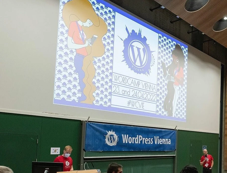 Projizierter Bildschirm zeigt WordPress Vienna; zwei Personen in roten Shirts stehen am Pult, Banner im Hintergrund.